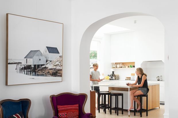 Bright contemporary interior view from a living area into a white kitchen through a large arched opening. The kitchen has a central island with a pale stone or light benchtop, timber-toned cabinetry, dark stools, open shelving, and integrated appliances. Two people sit and stand at the island. The foreground includes partial upholstered armchairs and a large framed coastal photograph, while a skylight or roof window adds natural light.