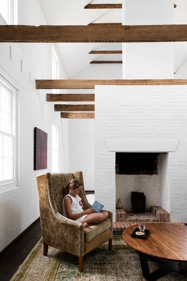 Bright, vertically framed living room in a renovated white-painted brick interior. Exposed dark timber ceiling beams span the room, with tall white-framed windows along the left wall and a prominent white brick fireplace at the rear. A textured upholstered armchair, patterned rug, and low round timber coffee table create a warm seating area, with a child reading in the chair.