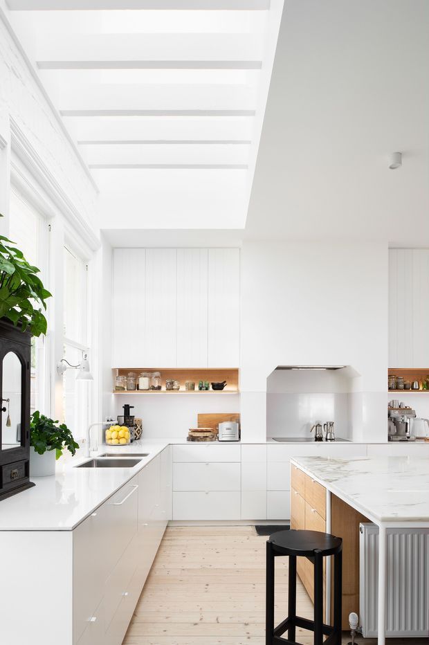 Bright white contemporary kitchen with extensive built-in cabinetry, pale timber floors, white stone benchtops, a central island with timber-panelled sides, integrated open shelving, and a stainless-steel cooking area. Large windows and exposed white ceiling beams create an airy, light-filled space. A black stool, dark freestanding stove, plants, and small appliances provide contrast.
