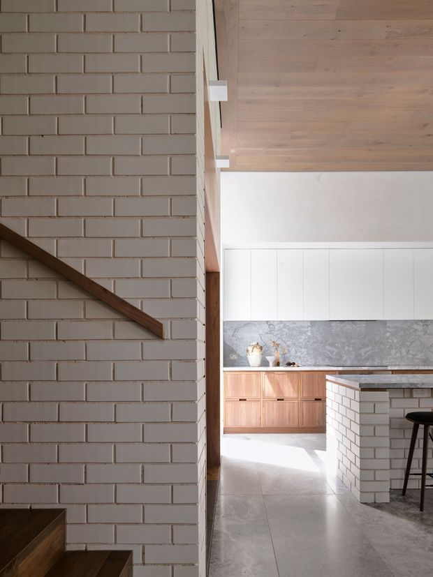 A refined contemporary kitchen viewed beside a stairway, featuring white painted brick walls, a timber stair handrail and timber-lined ceiling, white overhead cabinetry, warm timber lower cabinetry, a grey stone splashback and benchtop, and a white brick island with dark stools. The space is bright and materially textured, with a strong neutral palette and natural timber accents.