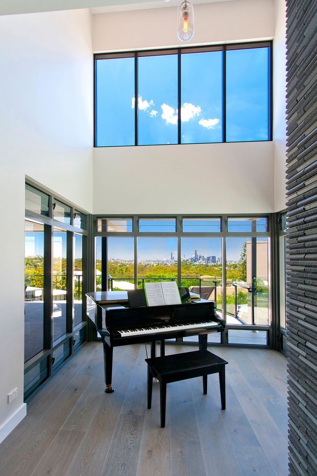 A bright double-height music room with a black grand piano and bench positioned in front of expansive black-framed glazing. The room features pale timber flooring, white walls, a textured dark feature wall, a suspended pendant light, and a city and leafy suburban outlook visible through the windows.