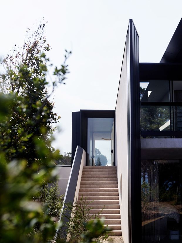 Exterior architectural photograph of a contemporary multi-level house, focused on a long straight staircase leading to a glazed entry. The building has crisp geometric forms, dark slender framing, pale rendered walls, extensive glazing, and a projecting upper-level volume. Mature landscaping partially screens the facade.