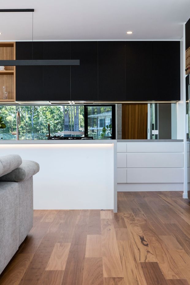 Contemporary open-plan kitchen viewed from an adjacent living area, with a large white island, pale timber flooring, extensive black overhead cabinetry, integrated white drawers, a dark linear pendant light, and views through glazing to leafy trees outside. A portion of a light grey sofa is visible in the foreground.