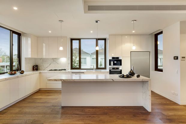 Bright, spacious contemporary kitchen with white full-height cabinetry, a large marble island with integrated seating, matching marble splashback, dark timber flooring, pendant lights, integrated appliances, and extensive timber-framed glazing. The open layout and neutral palette create a clean, refined atmosphere.