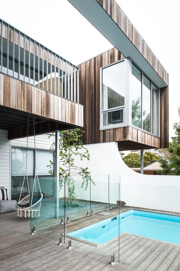 Exterior view of a contemporary multi-level residence organized around a small swimming pool and timber deck. The building features projecting upper volumes clad in vertical weathered timber, extensive aluminium-framed glazing, a glass balustrade, and a curved white masonry wall. A covered outdoor seating area with hanging chairs is visible beside the pool.