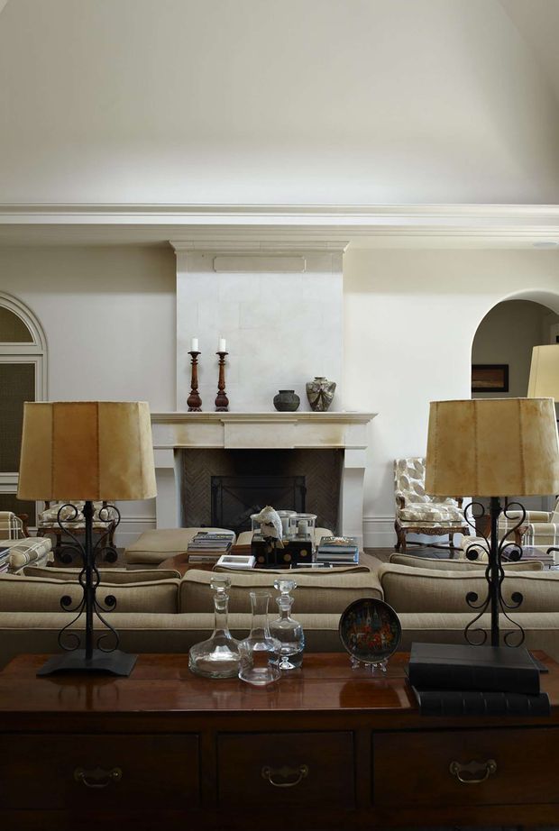 A formal, traditionally styled living room viewed across a dark wood console. The room features a symmetrical white-painted fireplace with classical mantel detailing and an inset dark firebox, upholstered beige seating, ornate wrought-iron and fabric lamps, decorative objects, and substantial ceiling cornicing. An arched opening and arched window detail reinforce the classic character.
