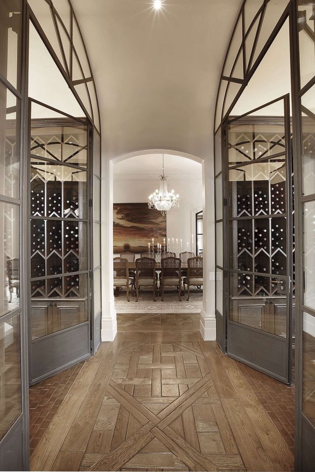 A refined interior view through tall, glazed metal wine-cellar doors into a formal dining room. The foreground features intricate patterned timber parquet flooring and dark-framed glass-and-metal wine storage cabinetry on both sides. Beyond an arched opening is a dining table with upholstered chairs, a crystal chandelier, candles, and large artwork. The palette is warm and neutral, with a traditional, elegant character.