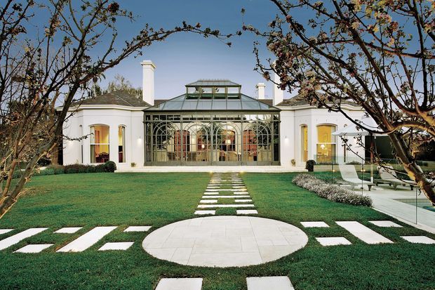A symmetrical, grand single-storey residence viewed from the landscaped front garden. The white rendered facade has classical detailing, tall arched windows, multiple chimneys, and a prominent glazed conservatory or atrium at the centre. A formal lawn is arranged with geometric white stepping stones and a circular paved feature, framed by mature trees.