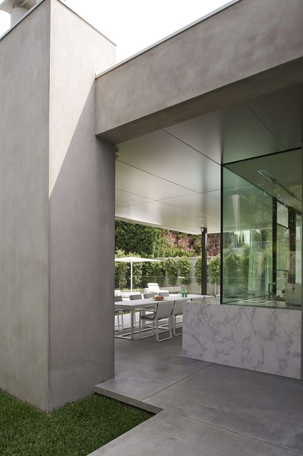A refined modern residential courtyard and outdoor entertaining area viewed from the exterior. Smooth pale rendered walls frame a deep roofed opening with extensive floor-to-ceiling glazing. Inside the opening is a marble-look stone counter or island, while beyond it is a covered dining terrace with a white outdoor dining table and chairs, surrounded by dense greenery. Large-format grey concrete-look paving, minimal detailing, and strong rectilinear geometry define the composition.
