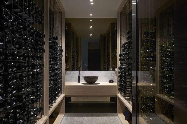 A dark, sophisticated wine room with extensive floor-to-ceiling bottle storage lining both sides of a central passage. At the end is a compact stone-topped wash or preparation counter with an undermount basin, gooseneck tap, open shelving, and recessed ceiling downlights. Glazed and metal-framed wine racks create reflective symmetry, while timber-toned cabinetry and a textured dark floor add warmth and depth.