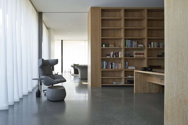 A refined open-plan interior featuring a built-in timber library and integrated desk, a dark sculptural lounge chair with ottoman, floor-to-ceiling sheer curtains, and a polished dark grey floor. The space is bright and minimalist, with a living area visible beyond the library partition.