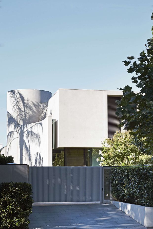 A contemporary multi-level residential facade viewed from the street, featuring smooth white rendered geometric volumes, a rounded curved wall, dark recessed glazing with horizontal louvres, and a tall corrugated metal front gate. A paved driveway, clipped hedges, raised white planter walls, and mature trees frame the entrance.