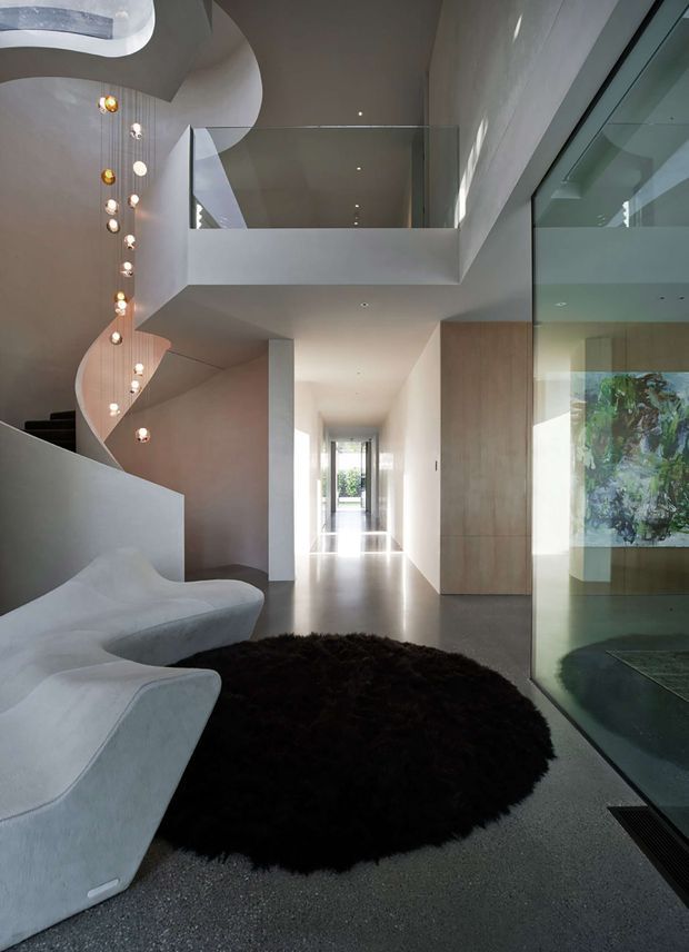 A sophisticated contemporary interior hallway and living area with a sculptural curved staircase, suspended decorative pendant lights, a flowing white built-in sofa, dark round rug, polished grey flooring, timber wall panelling, and extensive internal glazing. A mezzanine or upper-level void with a clear glass balustrade overlooks the space, while a long corridor leads toward bright glazing at the rear.