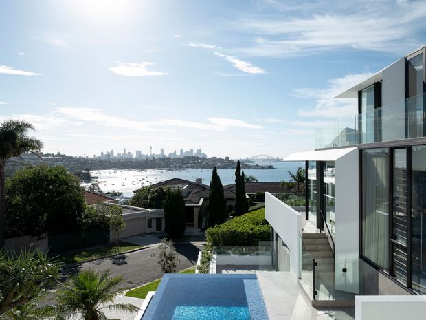 A contemporary multi-level residence overlooking a sparkling harbour, with a rooftop or upper-level terrace, glass balustrades, clean white rendered walls, dark vertical cladding, and a narrow swimming pool in the foreground. The elevated view includes a suburban neighbourhood, waterfront, and distant city skyline under a bright blue sky.