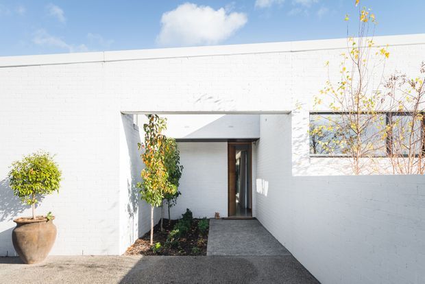 Bright minimalist exterior view of a white-painted brick facade forming a recessed entry courtyard. A covered rectangular passage leads to a dark timber-framed entry door, with a textured grey concrete path, white masonry walls, and sparse ornamental planting in large pots and garden beds. Clear blue sky and strong sunlight create crisp shadows.