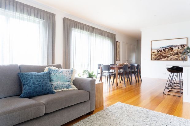 Bright open-plan living and dining area with a grey upholstered sofa in the foreground, large sheer-draped windows, timber flooring, a dark wood dining table with chairs, and a small kitchen or breakfast bar zone at right. The room has white walls, light natural finishes, and coastal-inspired blue textiles and artwork.