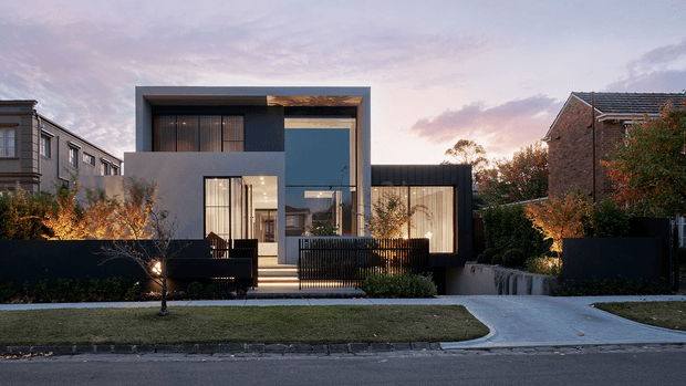 Contemporary two-storey urban residence viewed from across the street at dusk. The house has a strongly geometric composition with a dark charcoal and grey rendered facade, a projecting rectangular upper volume, extensive floor-to-ceiling glazing, and a lower wing with illuminated curtains. A front entry is reached by steps behind a dark vertical-slat fence, with landscaped planting, low retaining walls, lawn, and a concrete driveway. Traditional neighbouring homes frame the property.