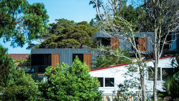 Exterior view of a contemporary multi-level residence partially screened by mature trees and planting. The building combines dark vertical cladding with projecting roof forms, warm timber louvre shutters, and a lighter lower-level facade with a red-trimmed roof edge.