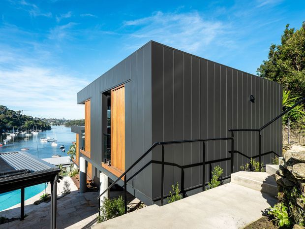 Exterior view of a compact modern multi-level building clad in dark vertical metal panels, with large timber-framed openings and a flat roof. Black metal balustrades line concrete steps and terraces, while a vivid waterfront harbour setting is visible beyond.