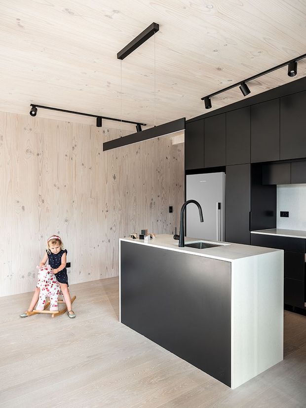 Contemporary open-plan kitchen with a large black-and-white island, integrated sink and matte black tap, full-height dark cabinetry, stainless-steel refrigerator, pale timber wall and ceiling panelling, and light timber flooring. Black track lighting and a linear pendant add contrast. A child on a rocking toy is visible at the side of the room.