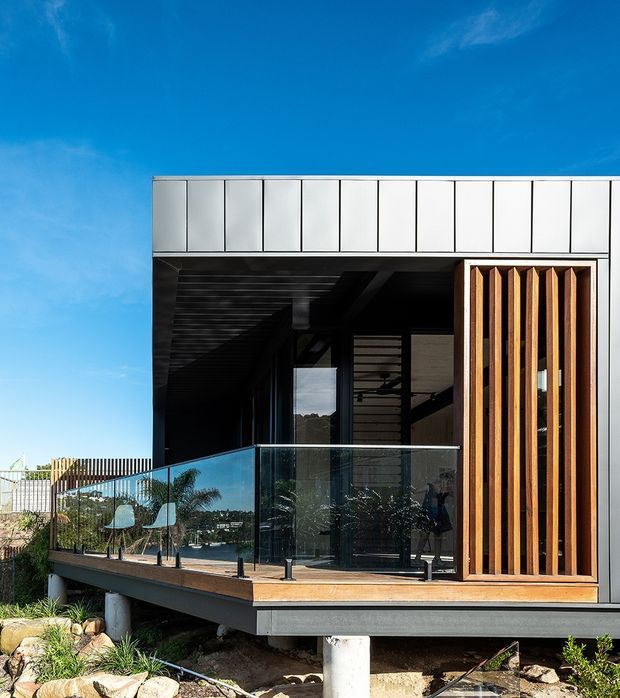 Contemporary elevated house exterior with a projecting balcony or terrace, transparent glass balustrade, dark recessed outdoor living area, and a striking vertical timber screen. The facade combines pale metal cladding with dark framing and timber detailing, set above a rocky landscaped site under a clear blue sky.