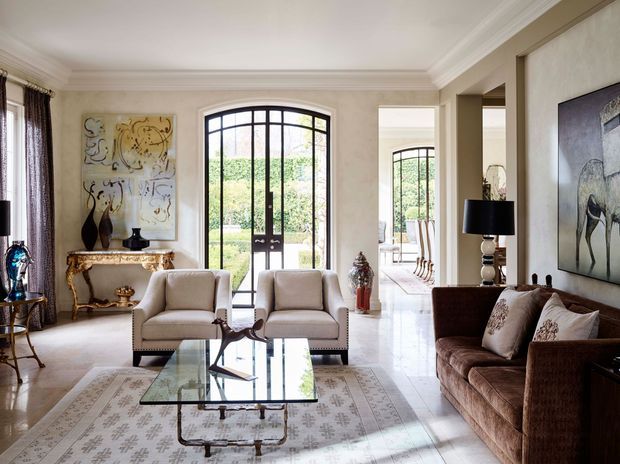 Elegant formal living room with a cream and beige palette, ornate ceiling cornices, pale stone or marble-look flooring, arched black-framed glazed doors, and layered traditional furnishings. Two light upholstered armchairs face a glass coffee table, while a brown textured sofa sits to the right. A decorative console, artwork, lamps, and a connected dining area are visible through the opening. The room is bright, refined, and classically styled with contemporary black-framed glazing.
