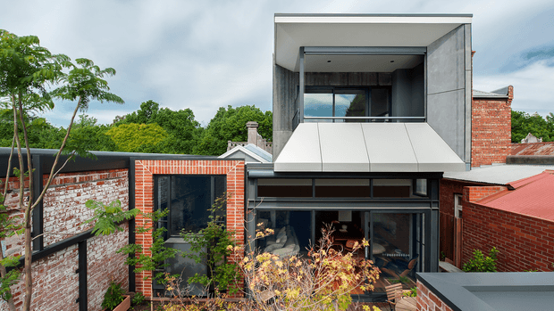 Exterior view of a contemporary multi-level residential extension combining original red brick masonry with a sharply defined upper volume. The upper storey features a deep recessed balcony or glazed opening, pale rendered or panelled walls, dark-framed glazing, and a faceted projecting white façade element. The lower level has extensive dark-framed glazing and opens toward a landscaped courtyard, while the adjoining original brick structures and pitched roof remain visible. The composition shows a strong contrast between heritage brickwork and modern geometric additions.