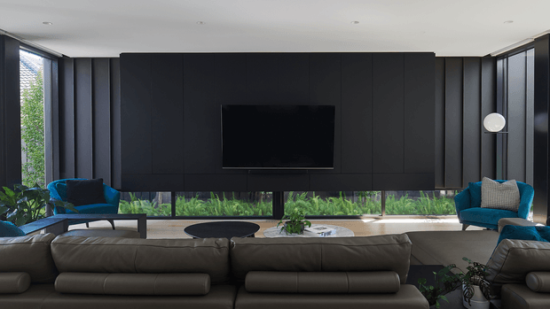 A sophisticated contemporary living room with a large dark built-in media wall and centrally mounted television. Floor-to-ceiling glazing wraps around the room, overlooking lush greenery, while a broad neutral sectional sofa faces the screen. Deep teal-blue lounge chairs, indoor plants, a low dark coffee table, and a slim globe floor lamp add contrast. The space has recessed ceiling lighting, dark vertical wall panelling, and a calm, refined atmosphere.
