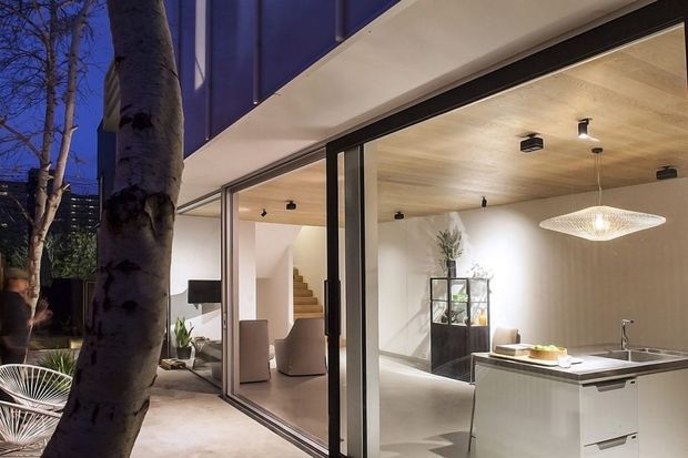 Evening view of a contemporary open-plan kitchen and living area through expansive floor-to-ceiling sliding glass doors. The interior has a pale timber-lined ceiling, white walls and cabinetry, a dark-edged kitchen island with integrated sink, a sculptural pendant light, and a visible stairway leading to an upper level. A paved outdoor area and tree are visible outside.