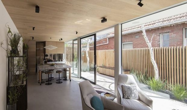 A bright contemporary open-plan kitchen and living area in a narrow extension, featuring a long island with bar seating, pale timber ceiling, polished light flooring, black track lighting, and full-height black-framed glazing opening to a paved courtyard. A compact seating area sits beside the glazing, with an exposed brick neighbouring wall and timber privacy fence visible outside.