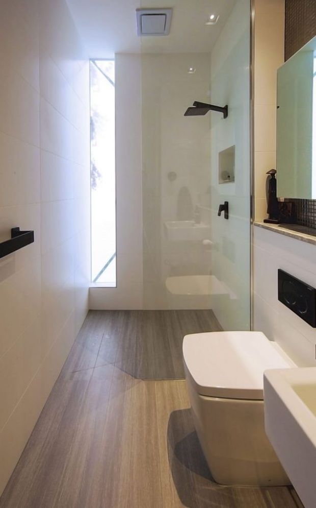 A narrow, contemporary bathroom with a walk-in glass shower at the rear, wall-mounted black shower fixtures, a wall-hung toilet, and a partial vanity with integrated basin at the right. Large light wall tiles, grey-brown wood-look floor tiles, recessed ceiling lights, a slim vertical window, and a dark mosaic backsplash create a clean modern fit-out.