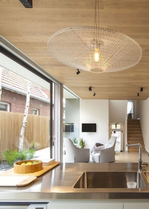 Bright open-plan kitchen and living area with a large stainless-steel sink set into a dark stone benchtop, white lower cabinetry, extensive floor-to-ceiling glazing, and a timber-lined ceiling. The adjoining living room contains pale upholstered chairs, a wall-mounted television, and a timber stairway. A large woven pendant light and black track lighting add focal details, while an outdoor courtyard is visible through the glazing.