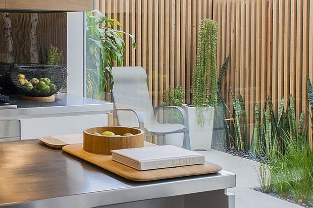A contemporary kitchen or dining space with a broad dark timber island or counter in the foreground, pale integrated cabinetry, and a light stone or composite work surface. A large glazed opening reflects an adjacent seating area, while a vertical timber slat wall and abundant potted greenery create a warm indoor-outdoor feel. The restrained palette combines timber, white, grey, and green with clean modern detailing.