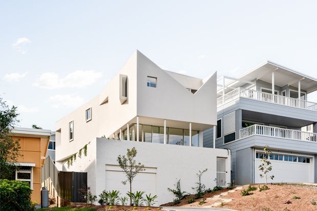 Exterior view of a contemporary multi-level residential building on a sloped site. The white rendered house has angular sculptural roof and wall forms, extensive glazing, a garage, landscaped planting, and a neighbouring or connected grey building with broad balconies and white balustrades.