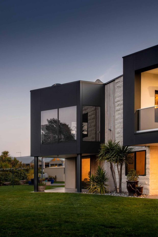 Twilight exterior view of a contemporary two-storey residence with bold black box-like volumes, extensive dark-framed glazing, pale textured masonry and vertical timber-look cladding. A raised upper-level wing is supported by slender dark columns, creating a covered outdoor area below. Warm interior lighting glows through the windows, with a manicured lawn, ornamental palms and neighbouring suburban homes visible around the house.