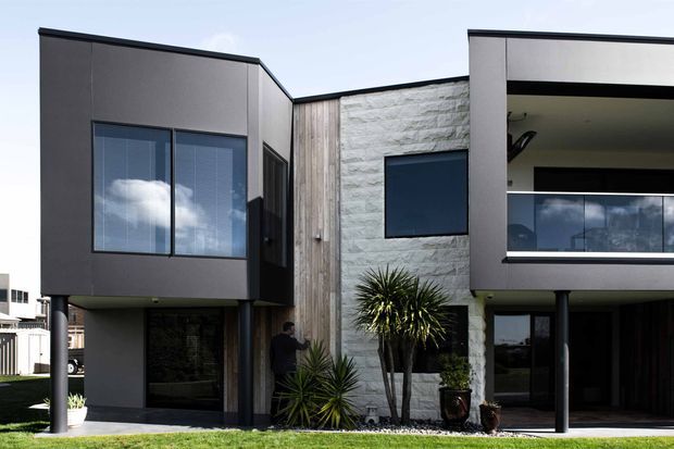 Street-facing view of a contemporary multi-storey residential building with strongly geometric, flat-roofed forms. The facade combines dark smooth panels, vertical timber-look cladding, a light textured masonry feature wall, extensive dark-framed glazing, and elevated upper-level volumes supported by slender columns. Minimal landscaping with sculptural yucca-like plants sits in front of the entry and covered outdoor area.