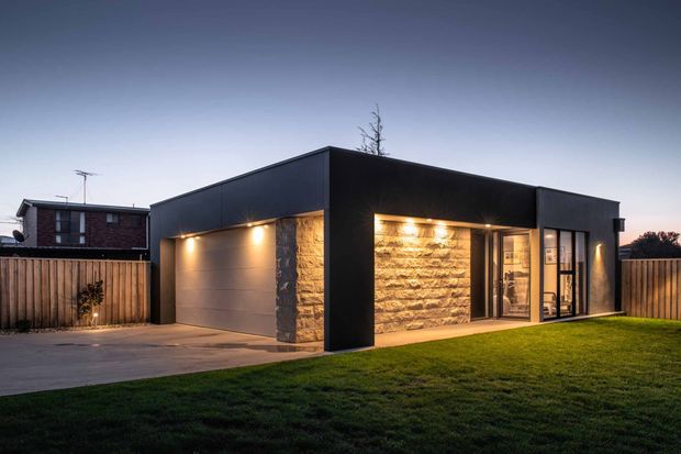 Contemporary single-storey home exterior at dusk, featuring a flat-roofed dark facade, integrated double garage, warm exterior lighting, large glazed entry doors, a prominent rough-cut stone feature wall, timber boundary fencing, paved driveway and lawn.