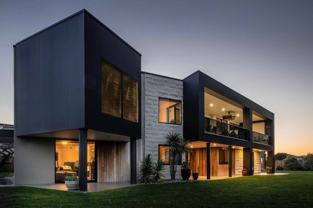 Contemporary multi-storey residence photographed at dusk, with a strong geometric composition of projecting dark-clad volumes, exposed structural supports, recessed glazing, warm timber entry and garage doors, and a central textured light-stone or concrete-block wall. The home includes a large illuminated upper-level covered balcony or terrace and extensive lawn landscaping.