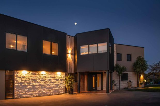 Twilight exterior photograph of a contemporary multi-level residence with strong geometric volumes, dark cladding, illuminated windows, a warm vertical timber feature, and a textured stone-clad lower facade. The recessed entry is framed by timber and glass doors, with integrated exterior lighting and minimal landscaped planting.