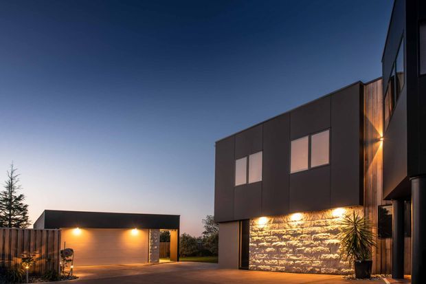 Exterior architectural photograph at dusk showing a contemporary multi-volume residence. The dark, rectilinear main facade has large square windows, vertical timber cladding, and a dramatically illuminated rough stone feature wall at ground level. A separate dark-roofed garage or ancillary structure sits to the left, connected by a paved driveway and landscaped garden elements. Warm exterior lighting contrasts with the cool blue evening sky.