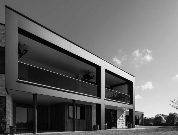 Black-and-white exterior photograph of a contemporary multi-storey residence with strong rectangular forms, deep recessed balconies, dark metal or glass balustrades, large glazed openings, and contrasting stone-textured wall sections. The house sits beside an open lawn and landscaped suburban setting under a partly cloudy sky.