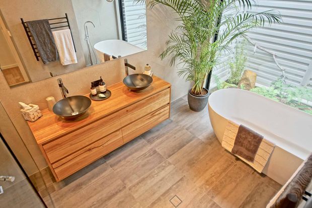Warm, contemporary bathroom with a double timber vanity, two vessel basins and wall-mounted taps, a freestanding white bathtub, large mirror, and indoor palm. The room features wood-look flooring, neutral textured walls, and a bright glazed or louvred opening overlooking greenery.