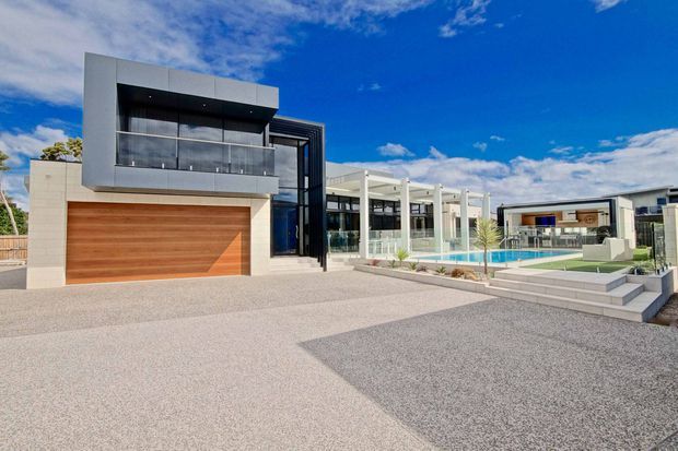 Wide exterior view of a large contemporary residence with a flat-roofed, geometric form. The home features a prominent timber-toned garage door, dark-framed upper-level glazing and balcony, extensive glass walls, a covered outdoor entertaining area, a swimming pool, paved terraces and landscaped areas. The palette is predominantly white, light grey and dark charcoal with warm timber accents under a bright blue sky.