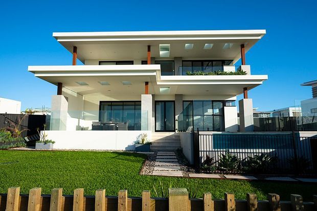 Contemporary multi-level residential facade with broad horizontal roof slabs, white rendered surfaces, extensive glazing, glass balustrades, timber-toned structural posts, and a landscaped front yard. A fenced swimming pool is visible beside the stepped entry, with neighboring modern homes in the background.