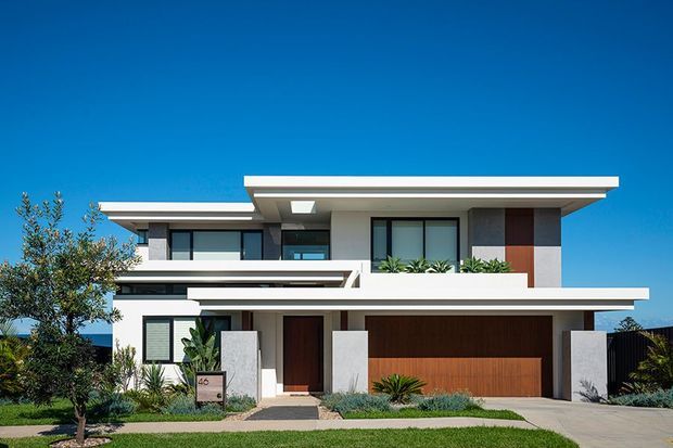 Front exterior view of a contemporary two-storey residence with strong horizontal lines, flat roof planes, projecting upper-level volumes, large dark-framed windows, rendered walls, and a wide timber-toned garage door. The landscaped frontage includes lawn, low planting, and a paved driveway under a clear blue sky.
