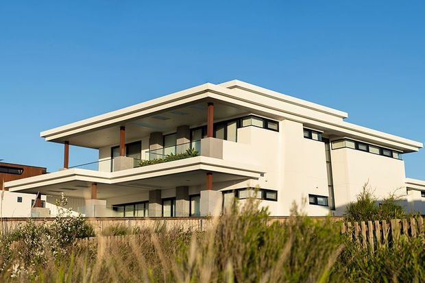 Exterior view of a large contemporary multi-storey residence with crisp white rendered walls, broad flat roof planes, projecting balconies, extensive dark-framed glazing, and slender timber-toned posts. The house sits behind coastal-looking grasses and a timber fence under a clear blue sky.