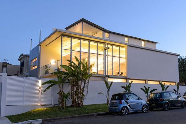 Contemporary multi-level residential home photographed at dusk, featuring a clean white rendered facade, extensive floor-to-ceiling glazing with horizontal blinds, projecting upper volumes, and warm interior lighting. A landscaped frontage with tropical planting, boundary walls, and parked cars is visible.