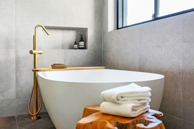 A refined modern bathroom featuring a freestanding white oval bathtub, large grey stone-look wall and floor tiles, a brushed gold floor-mounted bath filler, recessed wall niche with toiletries, and a high horizontal window. Folded white towels rest on a sculptural wooden stool in the foreground.