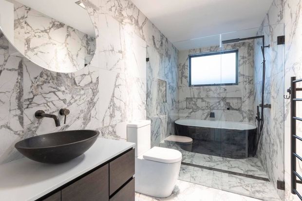 A bright, contemporary bathroom clad almost entirely in dramatic white-and-grey marble-look surfaces. The room features a dark vessel basin on a white benchtop with dark timber-look vanity cabinetry, a wall-mounted tap, a wall-hung toilet, and a freestanding bathtub within a tiled wet area. A black-framed shower and heated towel rail provide contrast, while a frosted window brings in natural light.