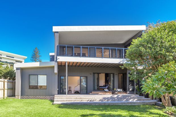 Exterior view of a contemporary two-storey residence with a flat roof, dark vertical cladding, white roof edges, extensive glazing, and a sheltered upper balcony. A broad timber-toned deck and lawn occupy the foreground, with mature landscaping framing the house.